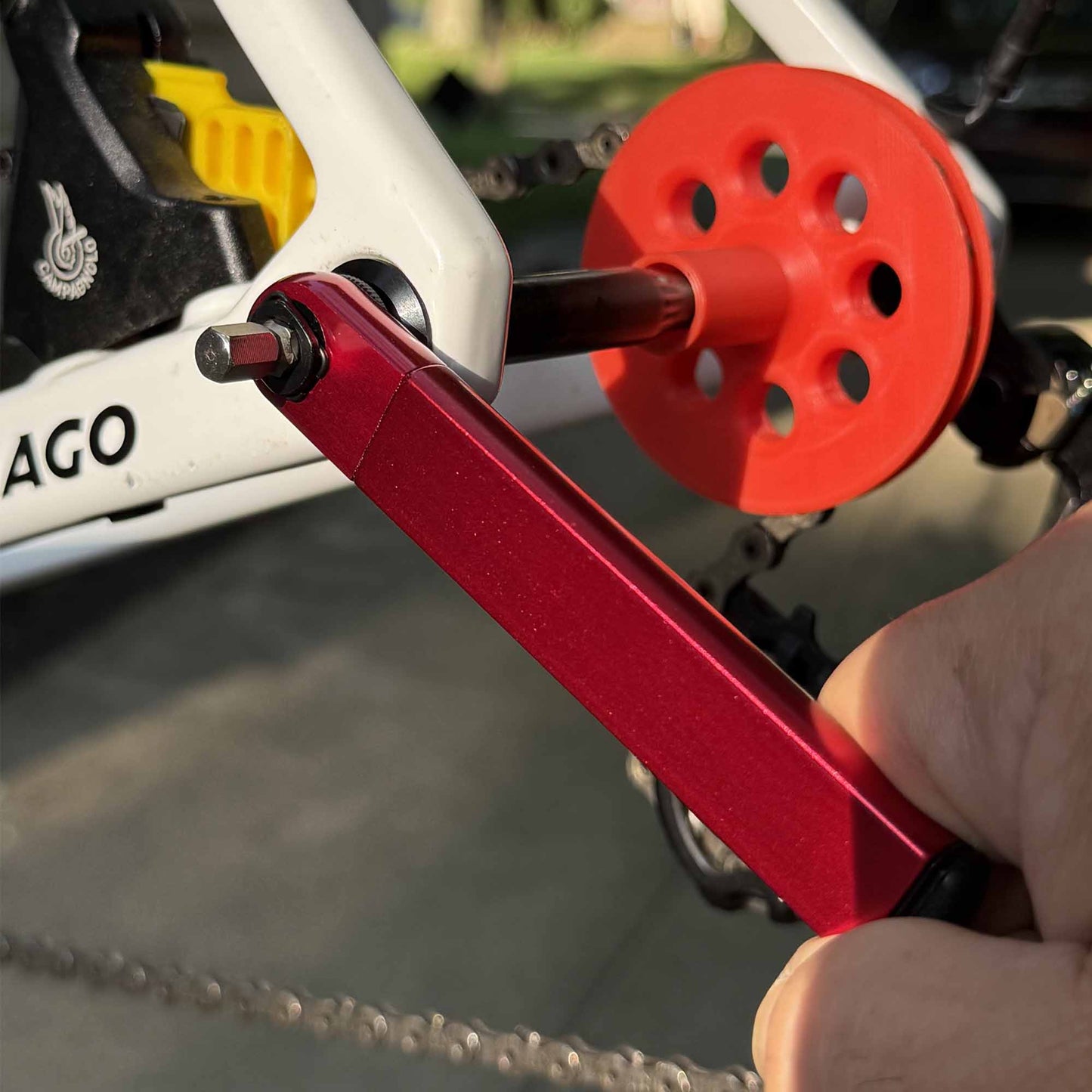 A hand uses the Prestacycle PocketRatchet Lite, a red multi-tool from Prestacycle, to adjust a bolt on a white bicycle frame. A red wheel and chain appear in the background.